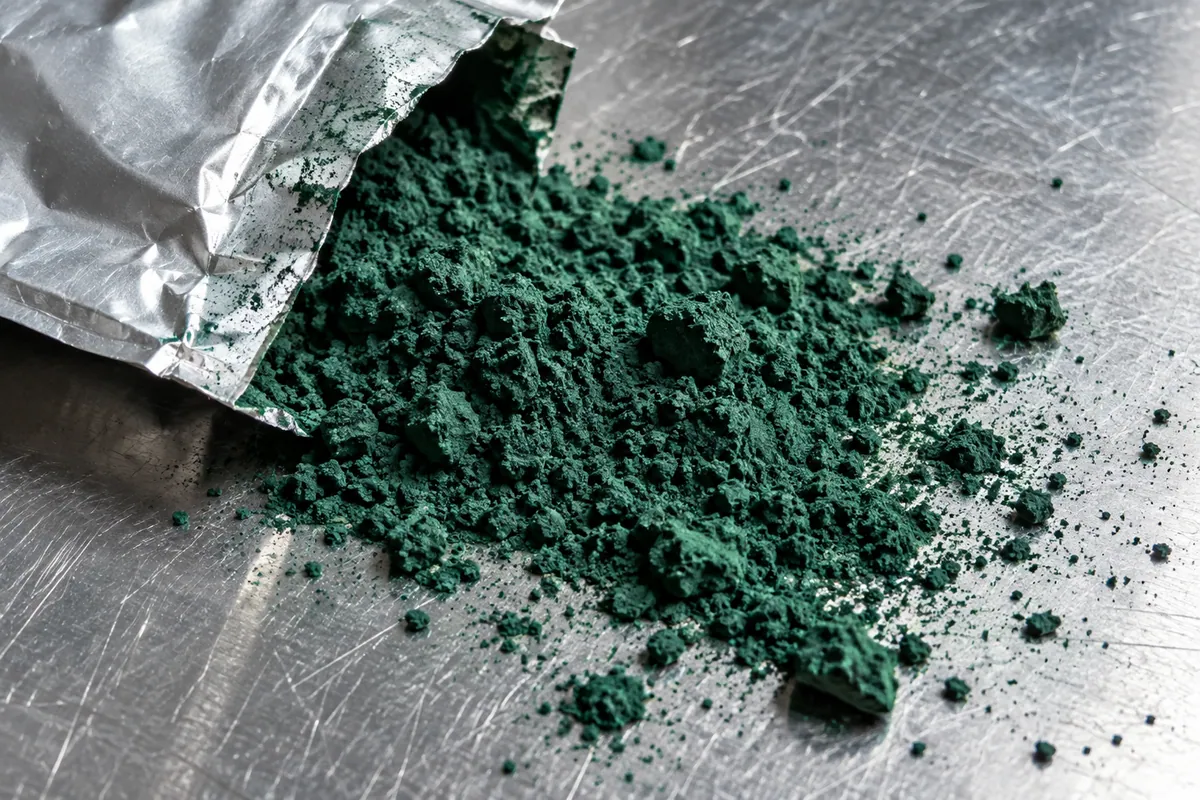 Sinivihreää spirulinajauhetta foliopussin reunalla teräspinnalla.